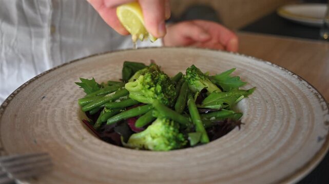 Hand squeezing fresh lemon over green vegetable salad with broccoli and beans