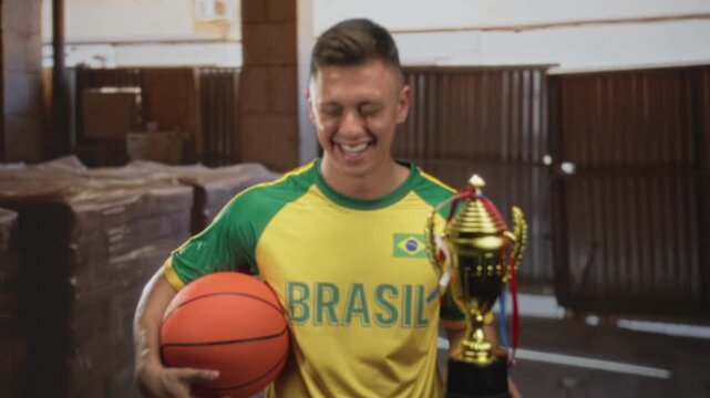 Man holding basketball and trophy in building wearing yellow green brasil jersey smiling broadly; victory joy pride.