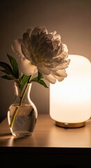 A white peony flower sits in a glass vase on a wooden table next to a glowing warm lamp in a cozy indoor setting at night.