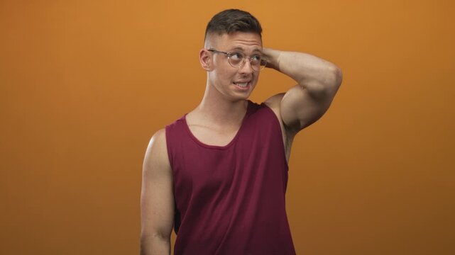 Man in burgundy tank top with glasses scratches back of head showing bicep and exposed skin in studio; awkwardness uncertainty.