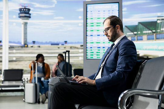 Entrepreneur traveling, international business trip, sitting in airport lounge, with baggage and laptop open. Businessman studies documents and plans while passengers move around, departure area.