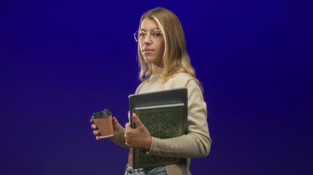 Young blonde woman wearing glasses and a beige sweater holds a coffee cup and clutches a book and tablet in studio; focus study routine.