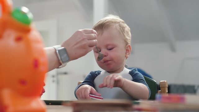 Caucasian baby spoon fed at table, parent hand feeding puree with spoon, orange toy blurred foreground, bib stained, baby tasting food, gentle caregiving in kitchen, soft natural light