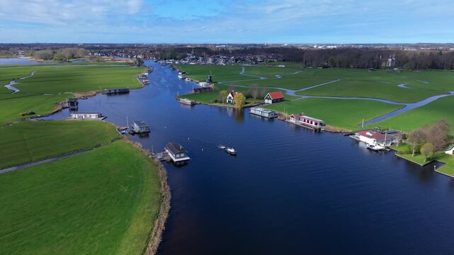 Aerial view of houseboats on a canal in the Netherlands. Scenic Dutch landscape with green polders, water channels, and traditional houses under a blue sky. Beautiful European village from above.
