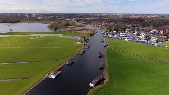 Aerial drone perspective of a Dutch town with a marina, traditional windmills, and modern houseboats along the river. Scenic landscape of the Netherlands featuring lush polders and waterways.