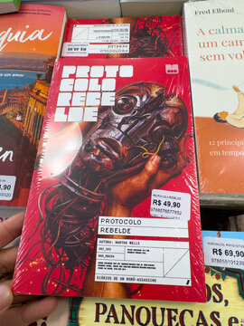 BELO HORIZONTE, MG, BRAZIL, MARCH 31, 2026. A dark, futuristic book cover for 'Protocolo Rebelde' with a robotic head design is held in a bookstore setting.