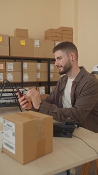 Man tapping smartphone with thumb beside parcel and stacked boxes at a packing desk in storage building; focus efficiency.