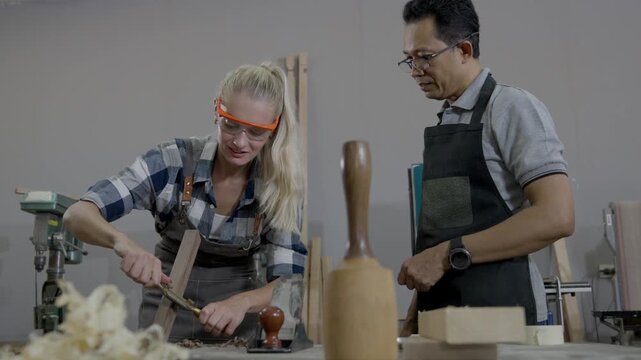 Woodworking workshop carpenter woman and man shaping wood with hand tool chisel and plane, teamwork training at workbench with safety glasses and apron, using tool in carpenter workshop with sawdust