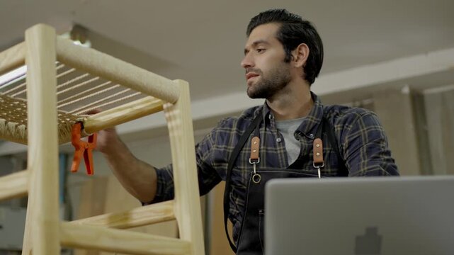 Carpenter woodworking craftsman in workshop planning handmade furniture chair project on laptop, using clamp on wooden frame, focused woodworker in indoor studio closeup casual maker