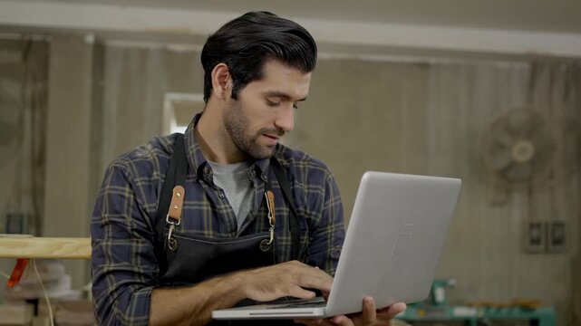 Man carpenter in woodworking workshop wearing apron using laptop for project planning and online orders, skilled craftsman with technology indoors portrait, focused furniture and tool studio work