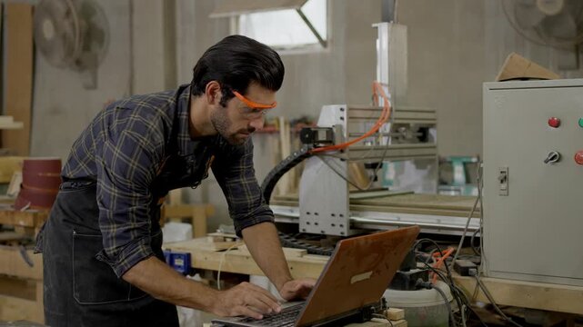 Male carpenter workshop woodworking with cnc machine workbench wearing safety glasses and apron, using tool and machine while checking equipment laptop, engineer technician worker focused fabrication