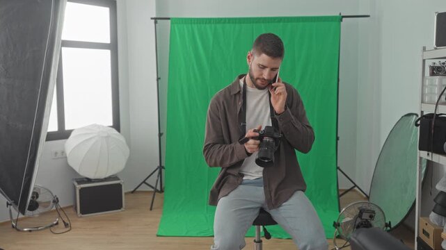 Man holding dslr camera and talking on smartphone while adjusting lens and checking settings against a green screen backdrop in a studio; concentration multitasking.