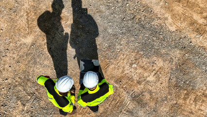 Aerial top down view of two engineers in safety gear discussing a project on a digital tablet. Construction workers standing on a dirt field casting long dark shadows in the bright sunlight. © ultramansk