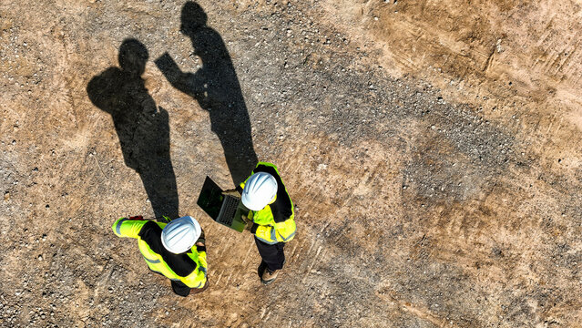 Aerial top down view of two engineers in safety gear discussing a project on a digital tablet. Construction workers standing on a dirt field casting long dark shadows in the bright sunlight.
