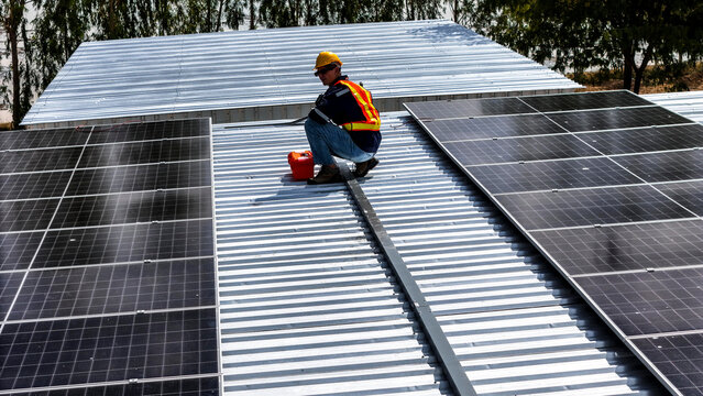 A worker wearing safety gear installing solar panels on a rooftop with nearby housing units in the background, emphasizing renewable energy, sustainability, and efficient solar power implementation