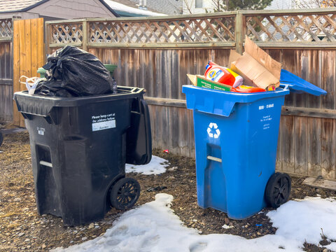 Calgary, Alberta, Canada. April 9, 2026. Overfilled black garbage and blue recycling bins stand in a snowy residential alleyway.