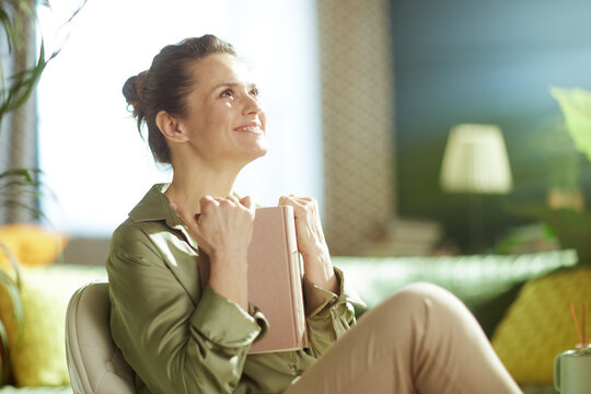 A woman pauses her reading to embrace a moment of inner peace and gratitude, holding her book close in a sunlit room filled with plants, reflecting on emotional well-being.