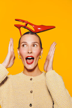 Surprised woman with red high heel shoe balanced on head looks excited with open mouth and raised hands. Bright yellow background and casual beige sweater create playful mood.