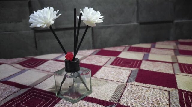 A close-up of an elegant reed diffuser with black sticks and white artificial flowers sitting on a patterned tablecloth. Ideal for home decor and aromatherapy concepts.