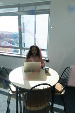African American woman sitting at round table by window, in pink blouse, using laptop, mug right