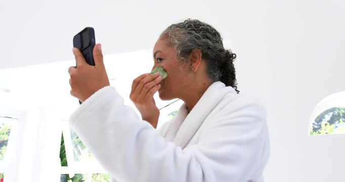 Senior woman in robe at home holding compact, checking mirror rubbing cheek with stone for skincare