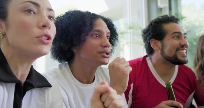 Diverse friends watching game leaning forward grimacing then calming red jersey with beer on couch