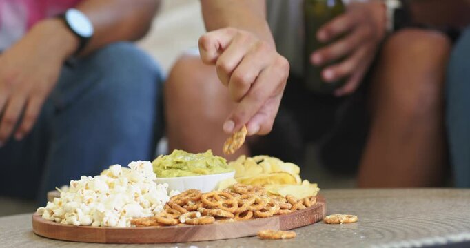 Diverse men grabbing snack on couch, causing curly man eating pretzel reacting to TV, green bottle