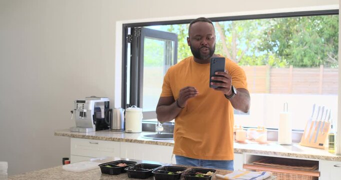 Mid adult African man checking phone, raising phone and filming four meal-prep boxes in kitchen