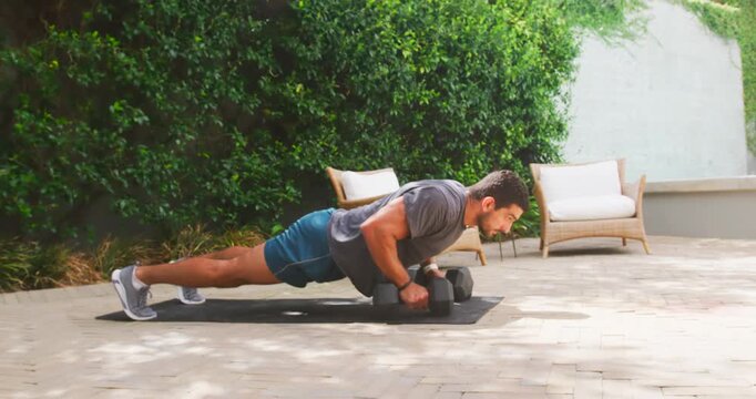 Man performing plank push-ups and single-arm hex dumbbell rows on patio mat, wearing sportswear