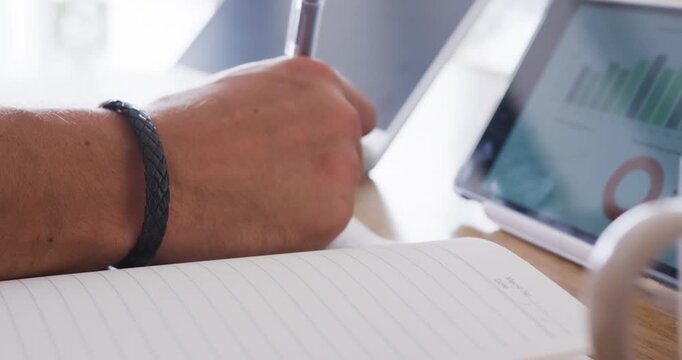 Man referencing tablet charts and writing notes at desk with pen in lined notebook for study