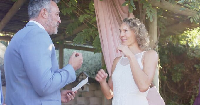 Man in light blue suit reading printed vows at wedding under wooden pergola, woman wiping tears