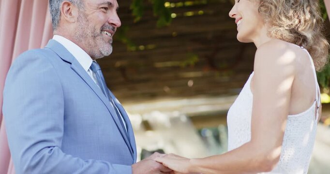 Couple holding hands while camera pulling back under pergola at wedding, man-in-suit woman-in-dress