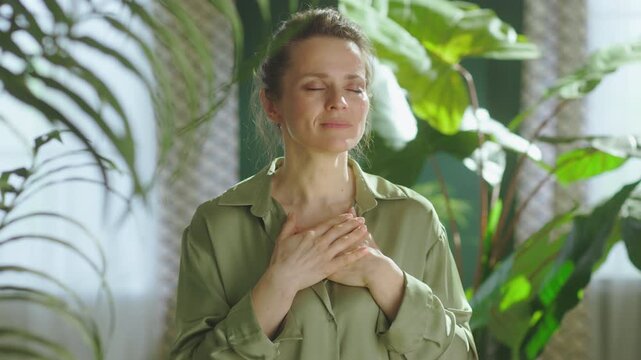 A serene woman stands with her eyes closed and hands over her heart, practicing deep breathing and mindfulness in a sunlit room filled with lush green plants.