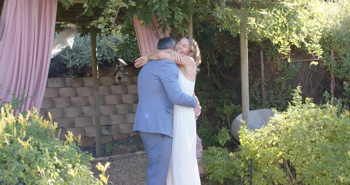Couple embracing under pink draped pergola, woman opening arms, celebrating in suit and white dress