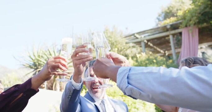 Diverse friends raising clear flutes after man in light blue suit cueing toast, clinking, sipping