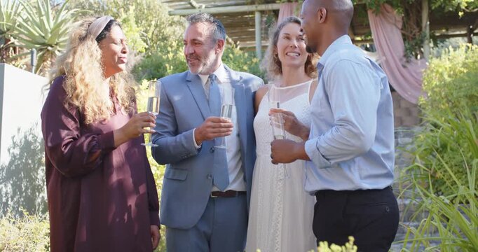 Diverse couple with two guests, woman in plum dress raising flute, others clinking, toasting garden