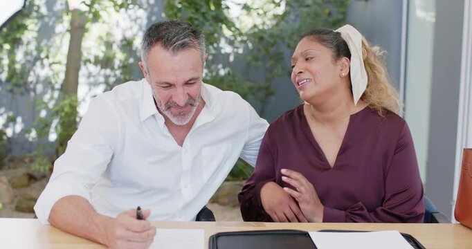 Diverse couple sitting at wooden table, man leaning in signing papers with pen, woman touching face