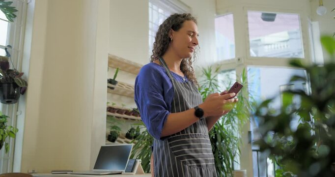 Non-binary adult in apron reading, tapping pink phone at plant shop, speaking, leaving voicemail