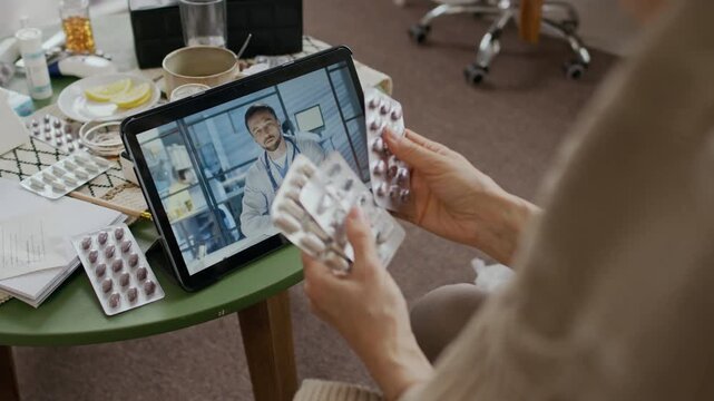 Over-shoulder shot of Caucasian male telemedicine doctor in white coat on tablet screen consulting unrecognizable senior female patient with flu, listening to complaints and advising on medications