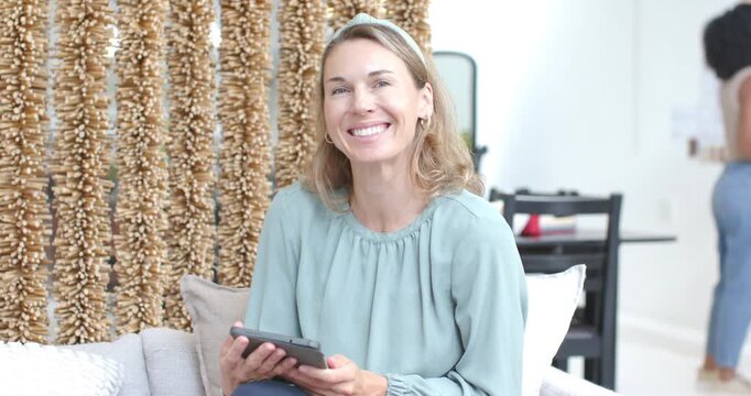 Vertical video: Smiling woman in seafoam top with tab at home laughing as pal entering, hanging art