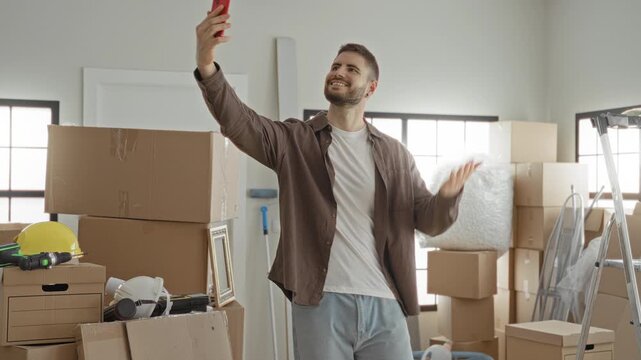 Man takes selfie with smartphone amid packed moving boxes, ladder, helmet and bubblewrap in building while unpacking and organizing; optimism new beginning.