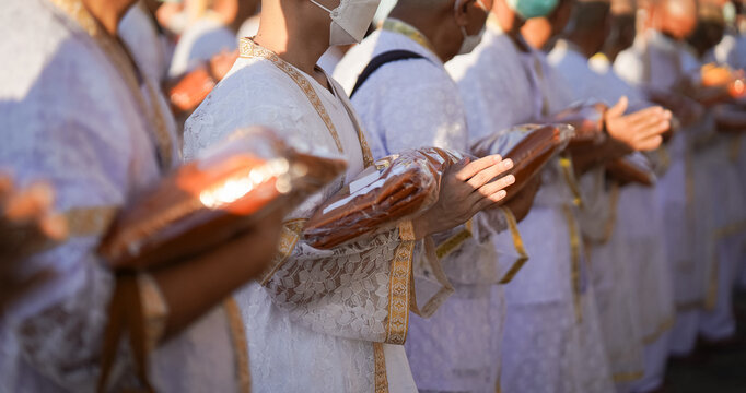 Thai men in white walk in line with palms together carrying folded orange monk robes as medium to large group approaches temple doorway, warm light before ordination.
