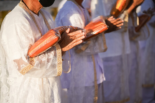 Thai men in white walk in line with palms together carrying folded orange monk robes as medium to large group approaches temple doorway, warm light before ordination.