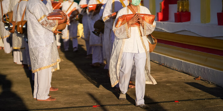 Thai men in white walk in line with palms together carrying folded orange monk robes as medium to large group approaches temple doorway, warm light before ordination.