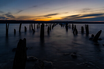 Obraz premium This tranquil landscape photo captures a serene shore at sunset. Old, weathered wooden pilings jut from calm waters, silhouetted against the bright sun and dark hills, evoking a peaceful mood.