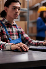 Fototapeta premium Young asian logistics manager analyzing goods supply on laptop. Warehouse worker doing inventory management on computer while sitting at desk with close up selective focus on hand