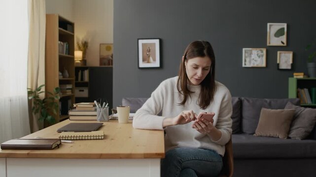 Medium full shot of young Caucasian woman sitting by work desk at home, browsing social media on smartphone or using lifestyle app, experiencing poor internet connection with frustrated reaction