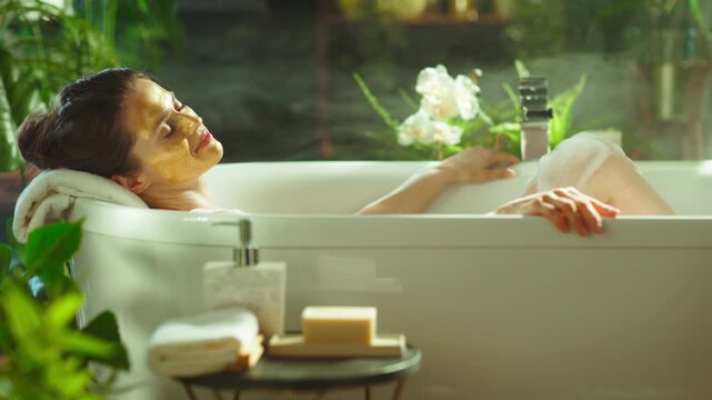 Serene, middle-aged woman with yellow facial mask relaxes in white bathtub amidst bubbles & steam. Lush plants & soft lighting create a tranquil spa setting for luxurious self-care & wellbeing.