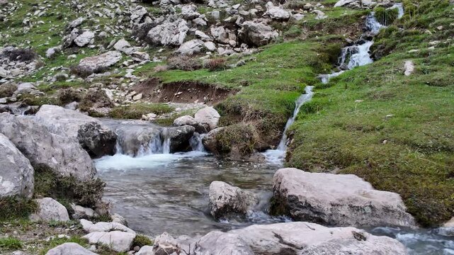 A scenic view of a clear mountain stream cascading down a rocky, grassy hillside, creating small waterfalls and pools in a natural alpine environment.