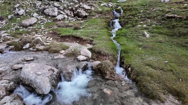 A small, clear mountain stream cascades down a rocky, green hillside, creating a series of gentle waterfalls as it flows into a larger pool below.